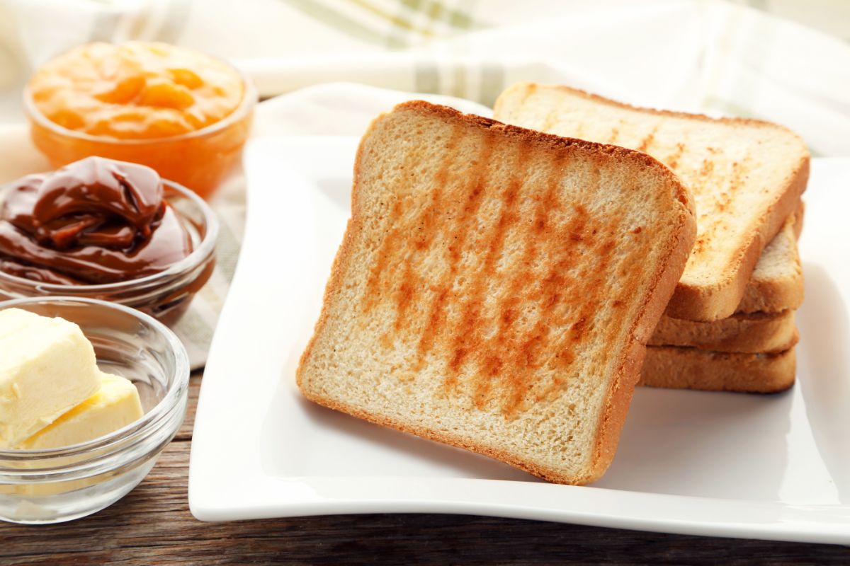 Toast bread slices on white plate next to small bowls of butter and jams