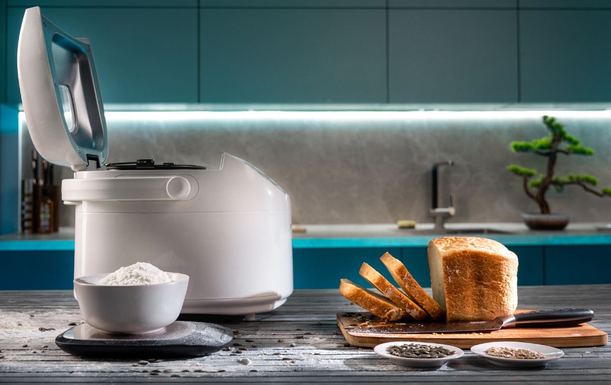 White breadmaker machine with bowl of flour on scale, plates with ingredients, loaf of bread on wooden pad with knife on kitchen table