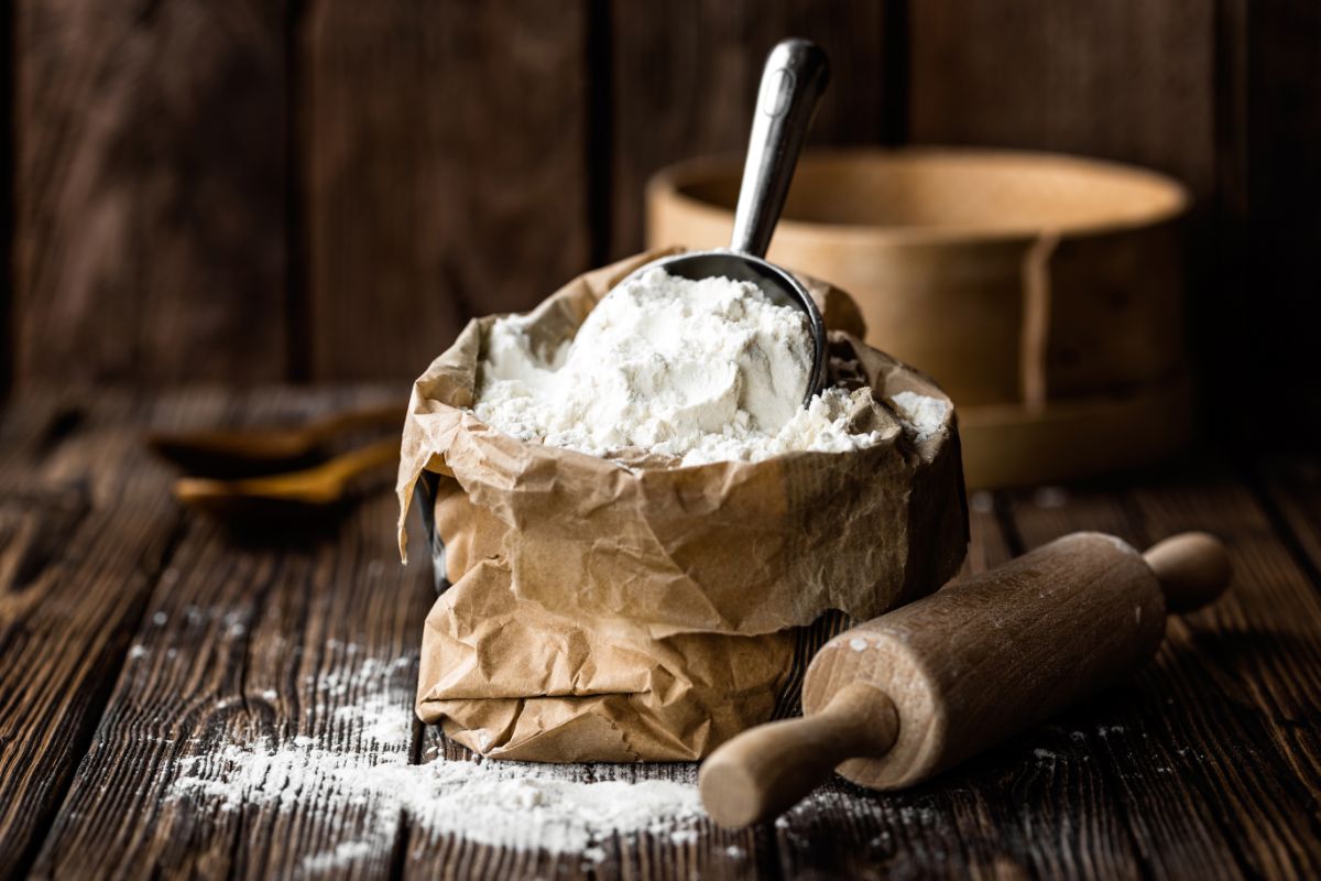 White flour in paper bag with metal spoon on wooden table with wooden kitchen roller and spilled flour