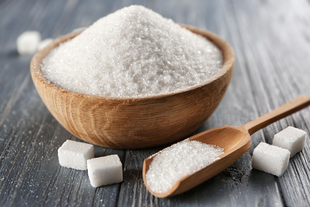 Wooden bowl full of sugar on black table with spoon of sugar and scattered cubes of sugar